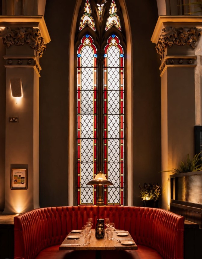 A red upholstered dining booth beneath a tall stained-glass window